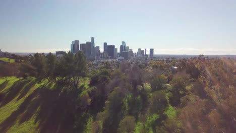 Vuela-Sobre-La-Colina-Cubierta-De-Hierba-Para-Revelar-El-Parque-Urbano-Y-El-Horizonte-Del-Centro-De-Los-Ángeles.