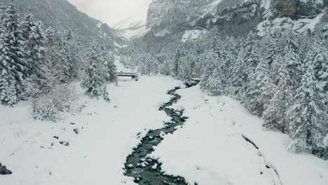 aerial-of-small-creek-running-through-snow-covered-landscape,-drone-slowly-lifting-up