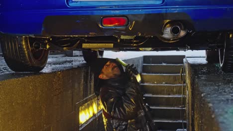 mechanic inspecting the underside of a car with a flashlight on a rainy day and with a raincoat on