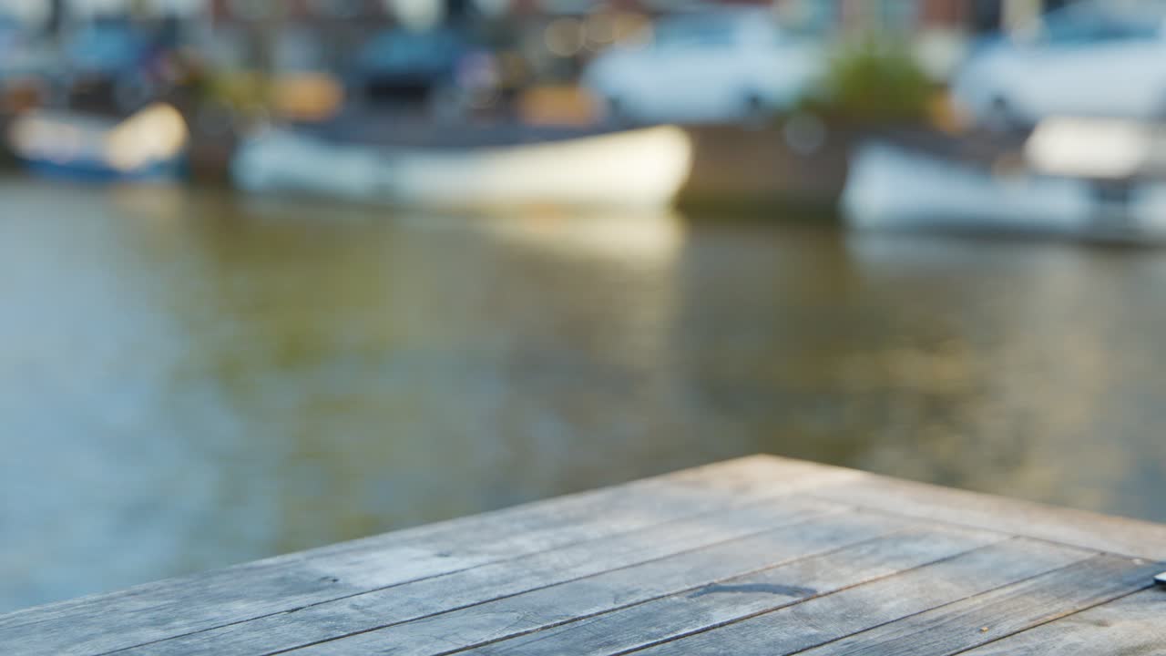 A hand sets down a pint glass filled with pale beer on a wooden table by a river, with soft natural daylight and blurred background