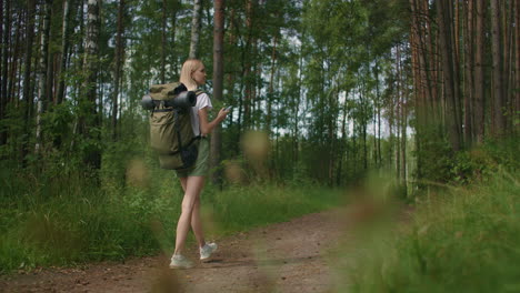 a young woman with a mobile phone walks through the forest traveling with a backpack in slow motion. navigate through the forest using the navigator in your mobile phone