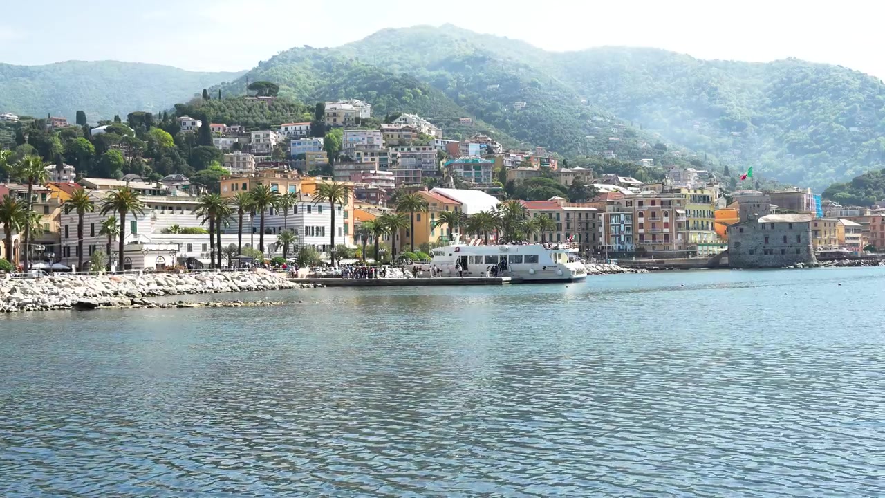 Scatto grandangolare del pittoresco porto di Rapallo, fiancheggiato da un'architettura vivace, palme e acque blu calme, con lo sfondo di rigogliose colline verdi sotto una morbida luce naturale.