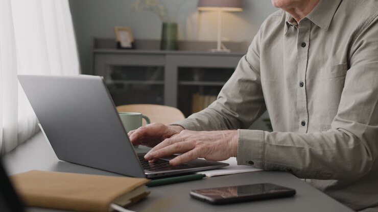 Camera Focuses On Elederly Man Hands Typewritting At Laptop And Holding Coffee Cup At Desk