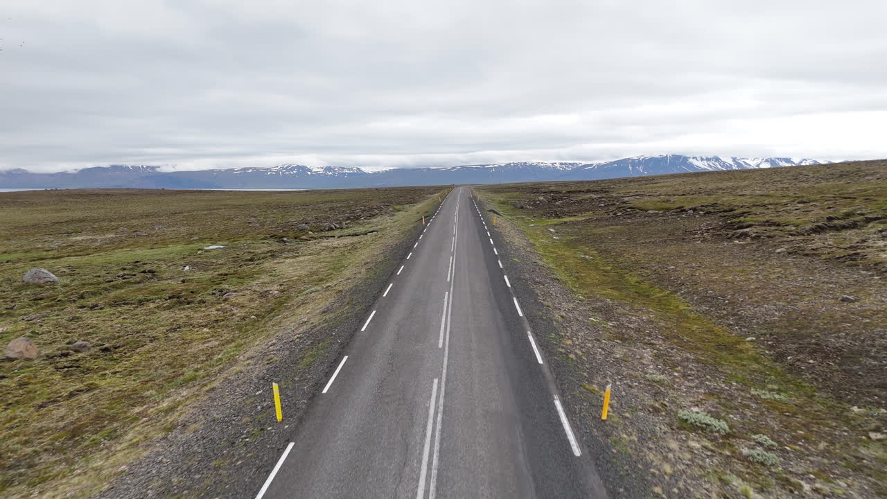 Dronebillede i fugleperspektiv af to cyklister, der kører langs en afsidesliggende kystvej i det østlige Island med fjorde, bjerge og åbent landskab under en overskyet himmel, hvilket symboliserer eventyrrejser, ensomhed og frihed.