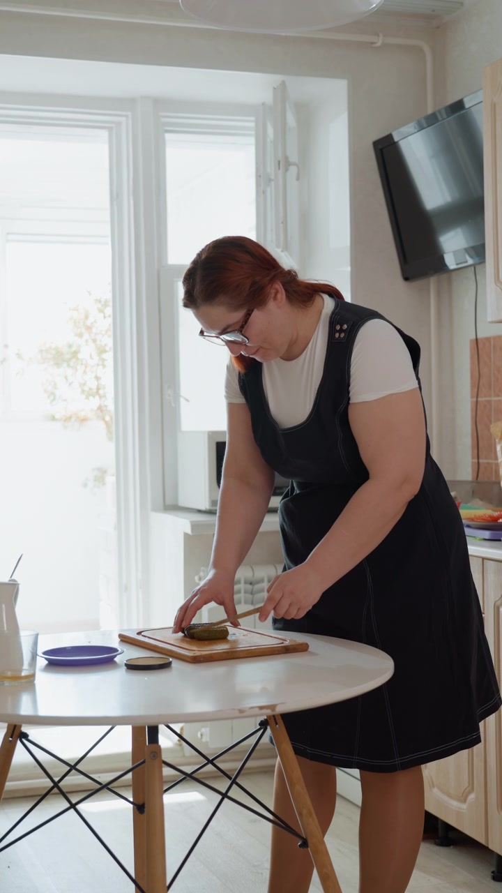 Ung kvinne står ved et rundt hvitt bord i et solfylt leilighetskjøkken, lener seg fremover for å smøre pålegg fra et glass på maten på et trefat, mens hun holder tallerkenen og strekker seg mot en kopp.