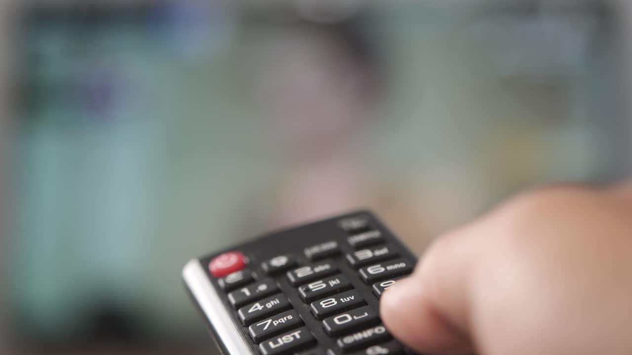 Person operating a TV remote control with a blurred television screen in the background