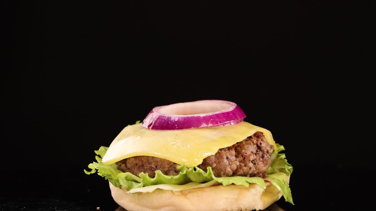 A slow-motion sequence shows a tomato slice and sesame seed bun top falling onto a beef burger with cheese, lettuce, and onion against a black background