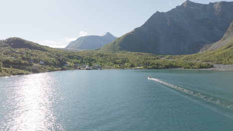 Boat-sailing-toward-waterfront-village-on-Norwegian-Lyngen-fjord-with-Scandinavian-Alps-in-background-on-sunny-summer-day,-Norway