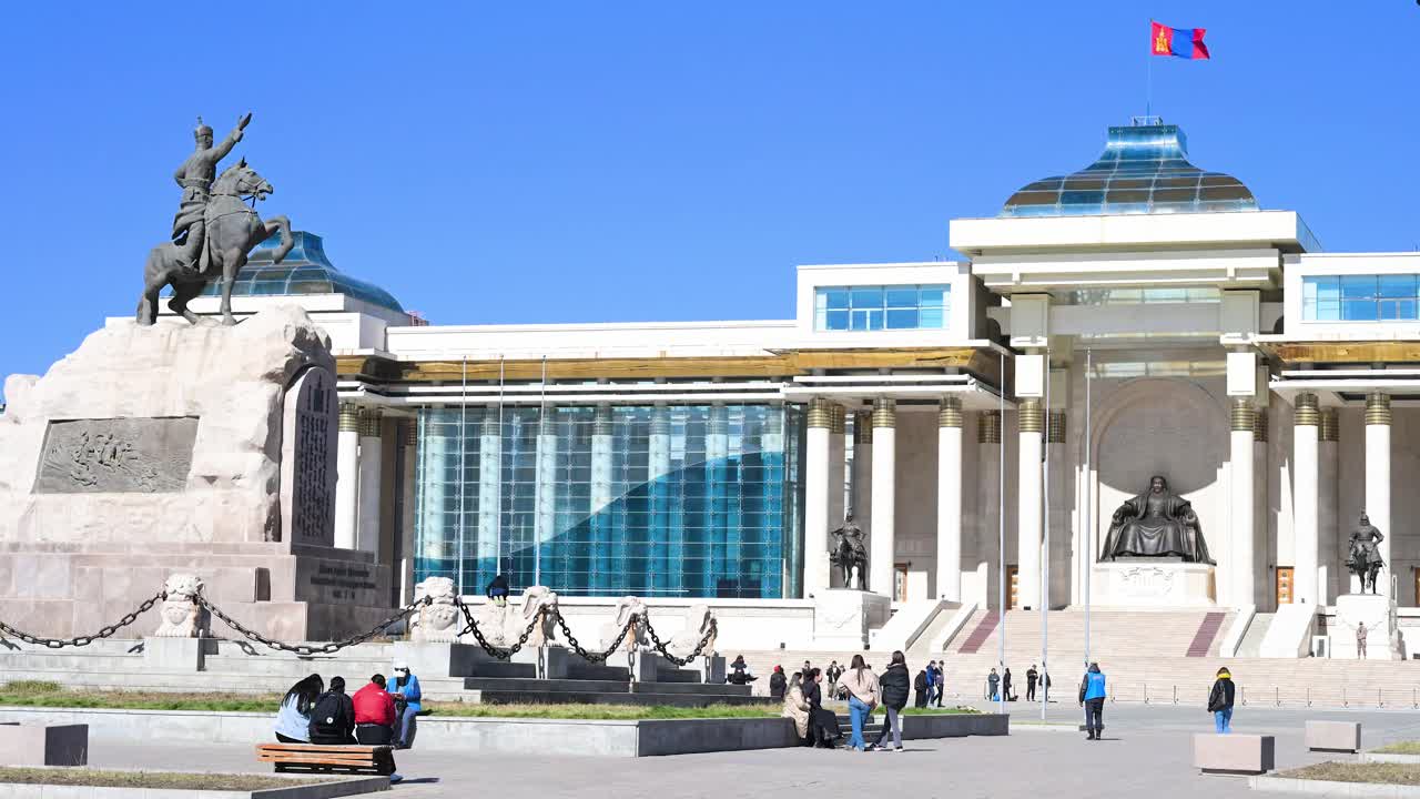On a sunny day, people gather at Sukhbaatar Square, with statues of Damdin Sukhbaatar (left) and Genghis Khan, the Government Palace in the background, and the Mongolian flag atop in Ulaanbaatar.