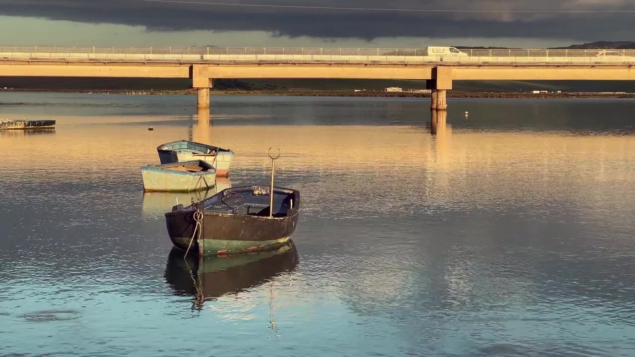 Gamle fiskerbåde flyder på roligt vand i Barbate, Andalusien, Spanien, ved solnedgang, med en bro i baggrunden og biler, der kører på den, hvilket skaber en fredfyldt og malerisk scene.