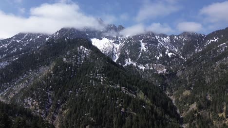 drone fly over forest with snow covered mountain range behind clouds in background
