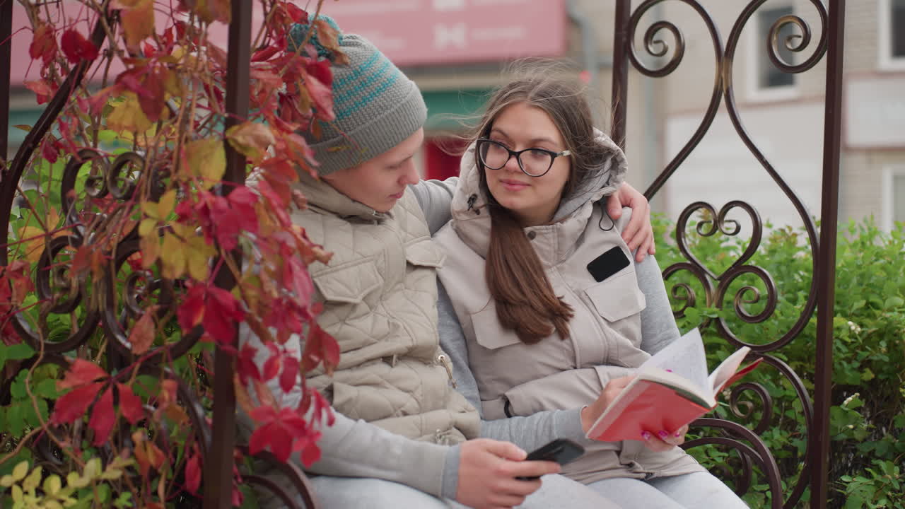 Lovely moment of lovers holding each other seated outdoor enjoying cool breeze as man warmly embraces woman reading book, surrounded by autumn leaves and greenery