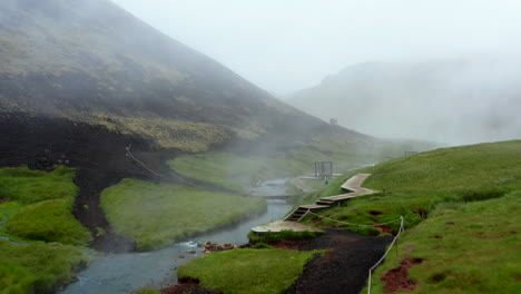 Vista-Aérea-Del-Valle-De-Reykjadalur-Con-Río-De-Aguas-Termales-Y-Piscina-Con-Exuberante-Pradera-De-Hierba-Verde-Y-Colinas-Con-Vapor-Geotérmico.-Drone-Vista-De-Bañarse-En-Aguas-Termales-Termales-En-Islandia