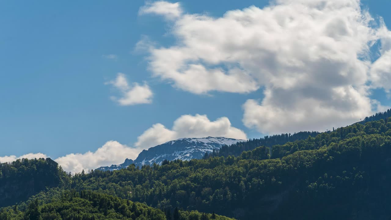 Timelapse on a spring day. Clouds swirl across the landscape with a snow-capped mountain in the background. The sun illuminates the mountainous landscape and its blooming nature.