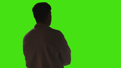 rear view of bored or fed up young man standing and waiting against green screen background with low key lighting