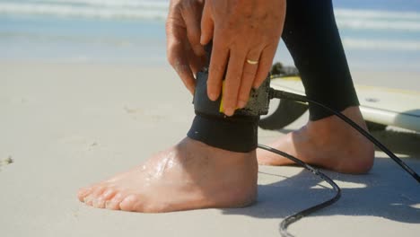 Low-section-of-active-senior-Caucasian-male-surfer-tying-surfboard-leash-on-her-leg-on-the-beach-4k