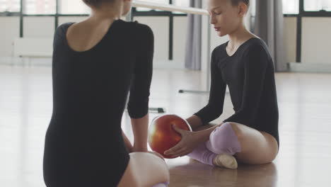 two gymnastic blonde girls talking while playing with a ball sitting on the floor before starting ballet class