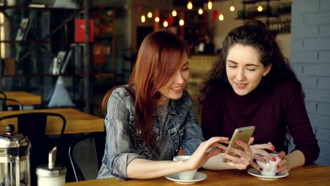 two young pretty girlfriends are using smartphone in cafe, laughing and talking emotionally. social media, meeting friends and modern technology concept.