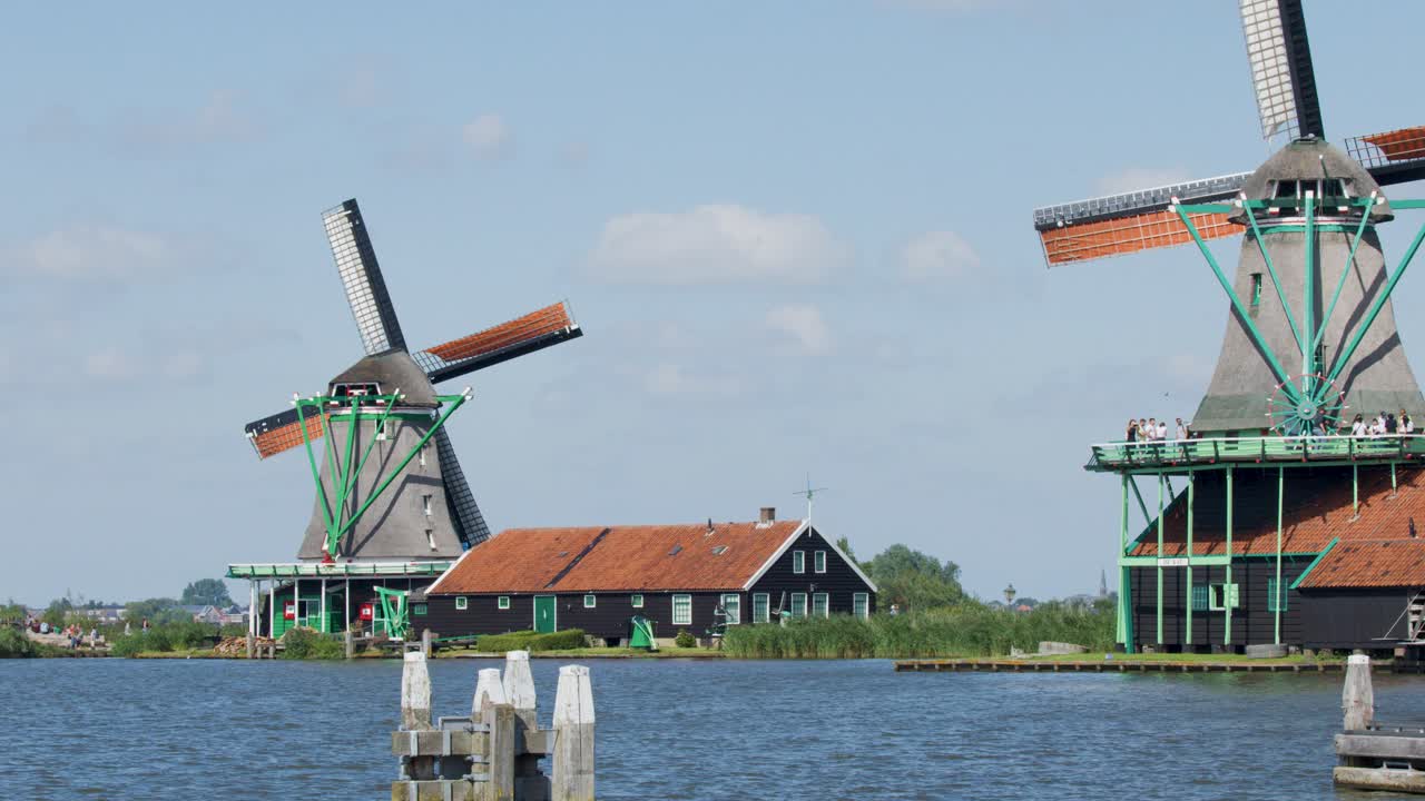 A classic Dutch windmill steadily rotates beside a calm river under bright daylight, with traditional houses and blue sky in Zaandam, Netherlands