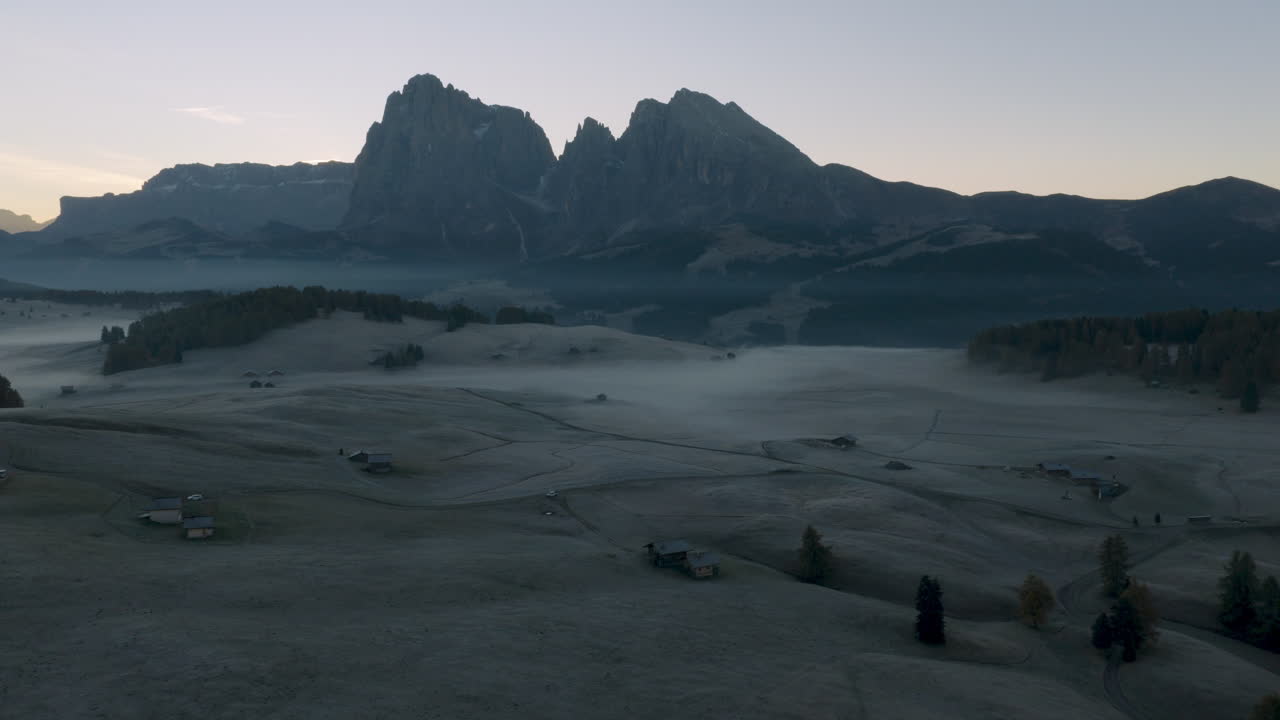 Cinematic aerial view of frosty Alpe di Siusi meadows and scattered mountain huts at dawn, mist drifting across rolling hills beneath the dramatic Sassolungo peaks in the Dolomites, Italian Alps
