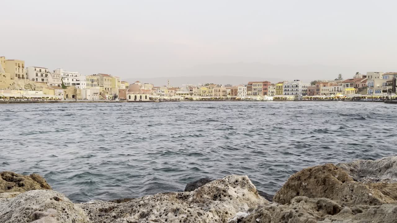 Slow reveal of Chania's Old Venetian Port, the main tourist area, with warm golden hour glow at sunset and hypnotising waves of the Cretan Sea [Chania, Crete, Greece]