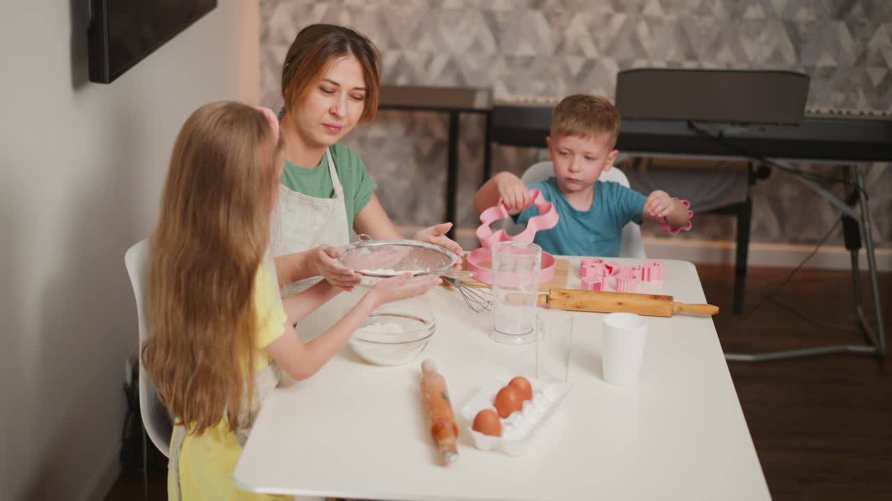 kid in yellow taps sieve to sift flour into bowl as baker holds handle with care, boy focused on toy shapes in background near piano, baking tools and eggs on kitchen table
