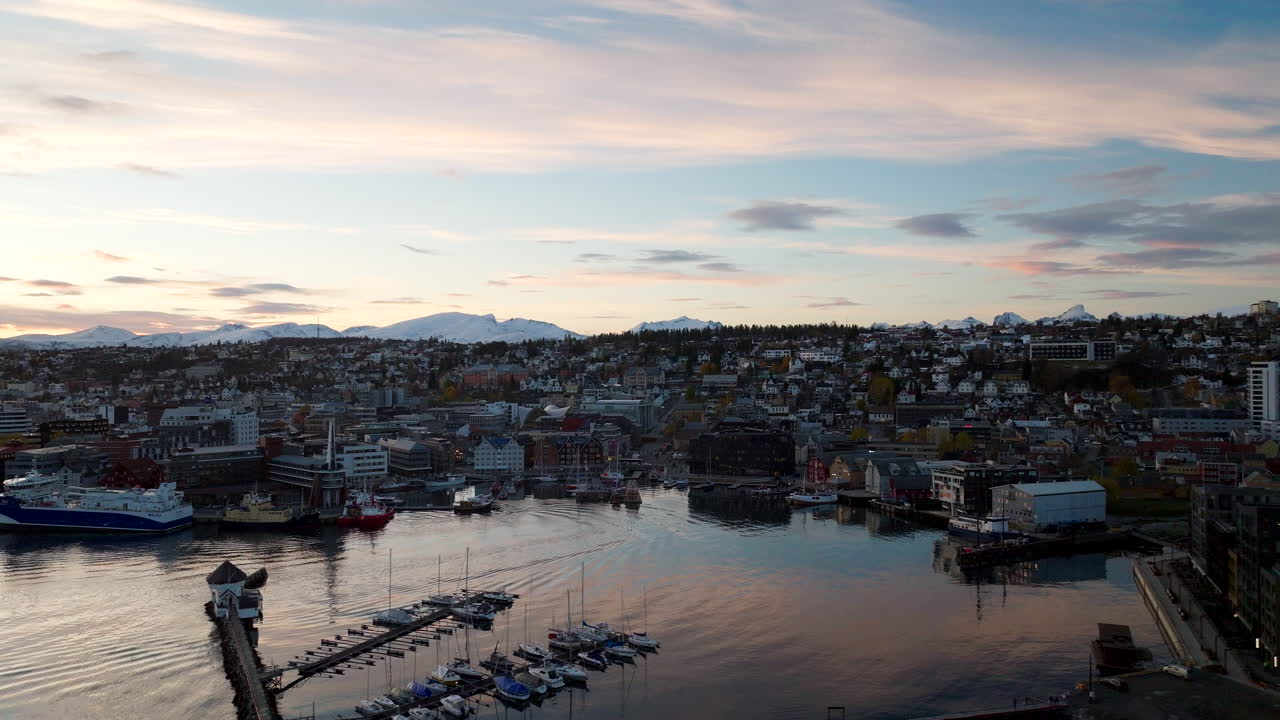 Aerial drone view of Tromso harbor during sunset in early winter. Light snow covers the mountains in the background while the city and boats reflect in the calm water of Northern Norway