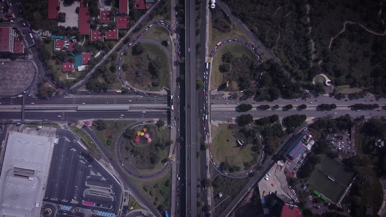 Overhead shot of cloverleaf intersection south of Mexico City, showing several cars driving, a parking lot, soccer fields in the lower right corner, trees and some red roofs