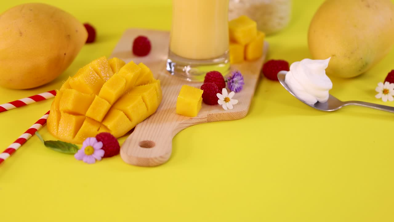 Glass of mango smoothie with cut mango, raspberries, whipped cream, and flowers on yellow background
