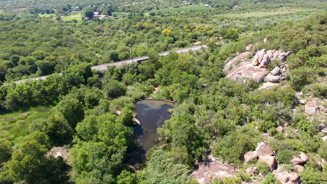 Small pond surrounded by green trees and grass near train track and small rocky mountains