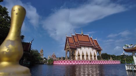 Estupa-Dorada-En-Primer-Plano-Con-Un-Templo-Tailandés-Tradicional-Junto-Al-Agua-Bajo-Un-Cielo-Azul