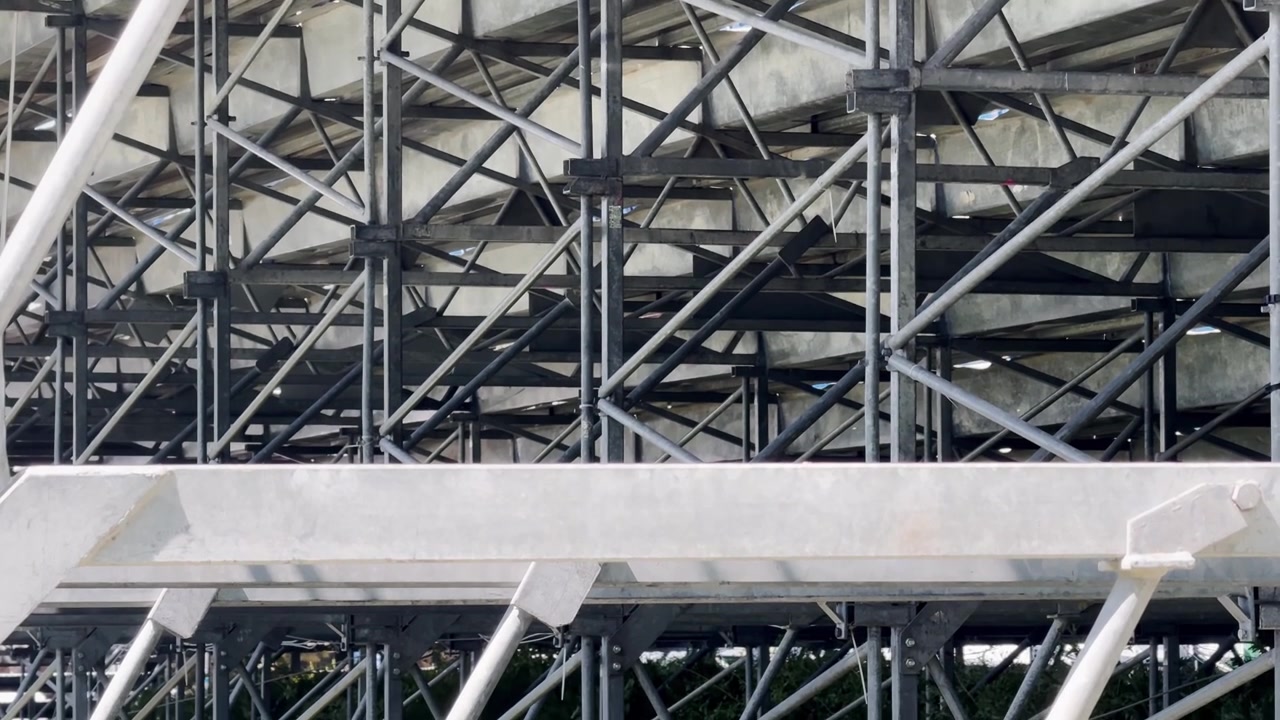 Parte inferiore delle tribune di un campo da tennis che mostra gradinate in cemento, travi di supporto in acciaio, controventature diagonali e la struttura di costruzione di un impianto sportivo.