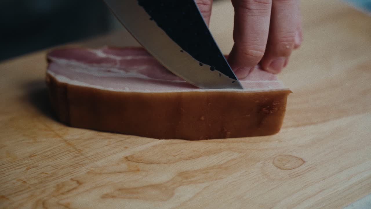un cuchillo afilado corta una pieza gruesa de jamón en una tabla de madera, mostrando los pasos de preparación de la comida
