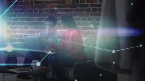 Animation-of-glowing-network-over-male-and-female-colleague-using-laptop-on-couch-in-office