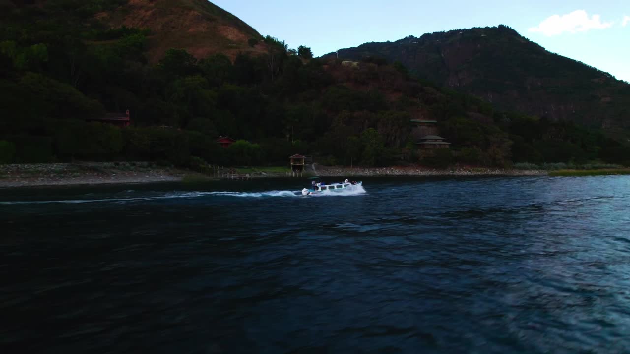 Panoramica aerea a destra che segue una barca che attraversa il Lago Atitlan durante il tramonto, con silhouette vulcaniche e riflessi colorati nel cielo