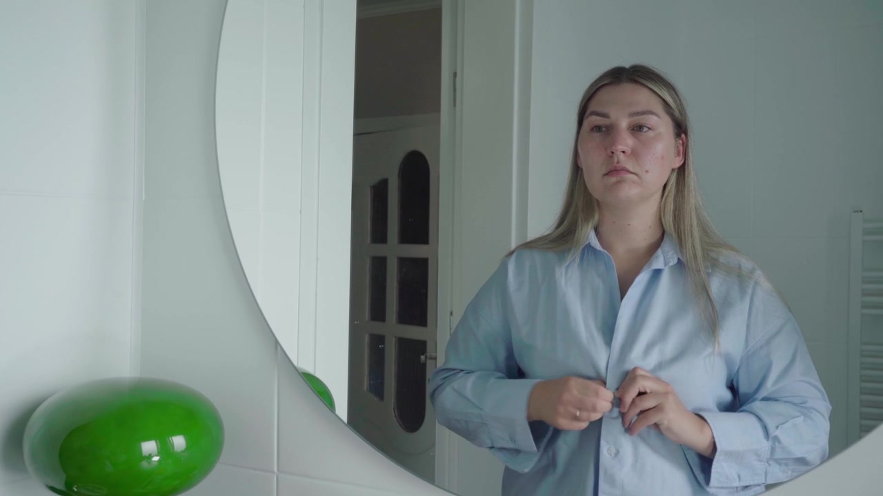 Una candidata si abbottona una camicia da lavoro chiara davanti a uno specchio, preparandosi prima di un colloquio di lavoro a distanza.