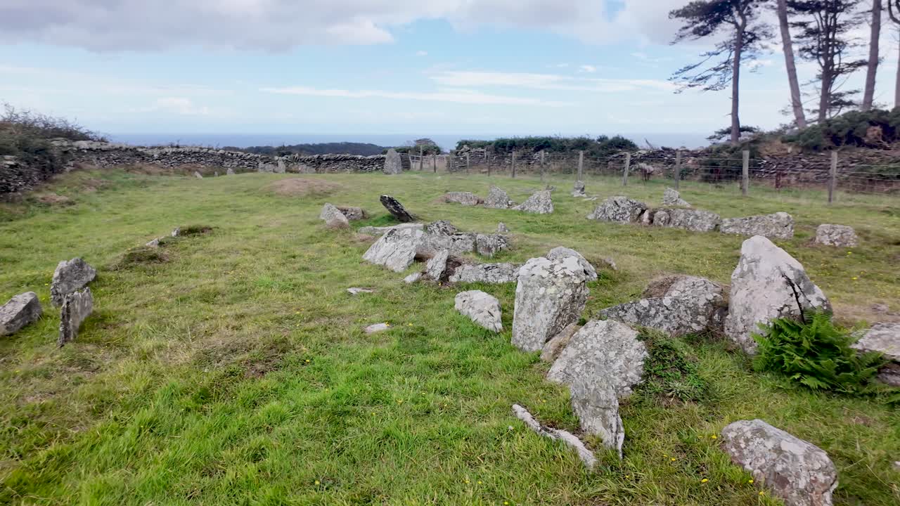Ancient stone tomb remains scattered across a grassy field with coastal views and trees in the Isle of Man.. Pan Left