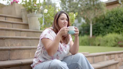 Happy-plus-size-biracial-woman-sitting-on-steps-in-garden-drinking-coffee,-copy-space,-slow-motion