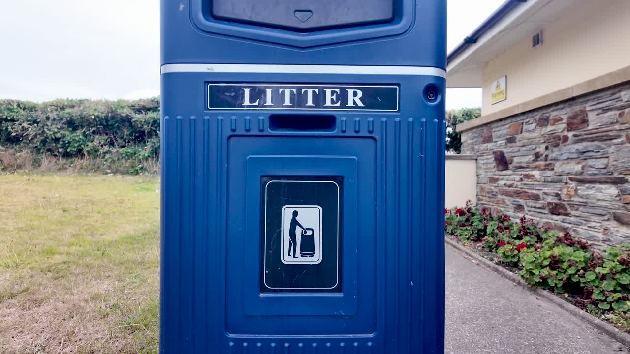 Blue public litter bin standing on grass in a park area beside a building