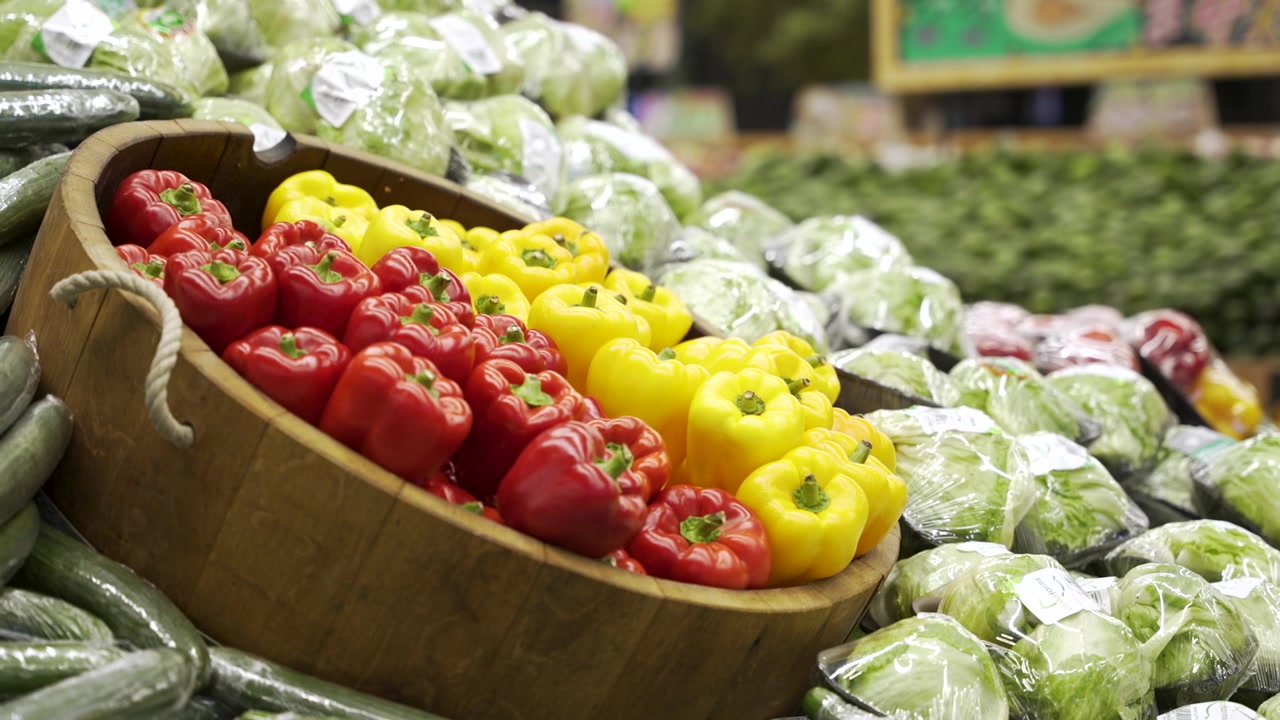 Aydınlık bir marketin sebze-meyve reyonunda, sarılı marul ve salatalıklarla çevrili ahşap bir sepeti dolduran kırmızı ve sarı dolmalık biberler.