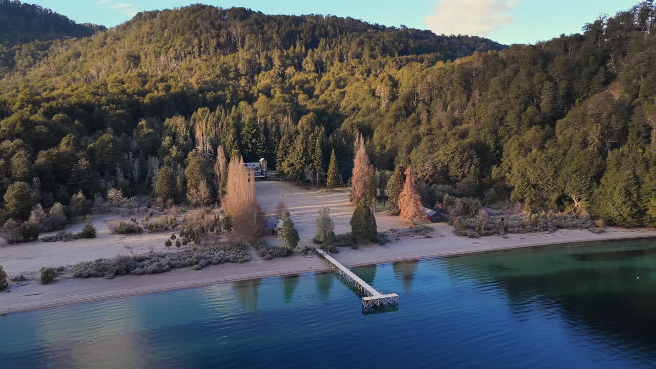 Piaszczyste wybrzeże jeziora z drewnianym molo i spokojną wodą obok zalesionych wzgórz nad Lago Espejo, Neuquén, Argentyna