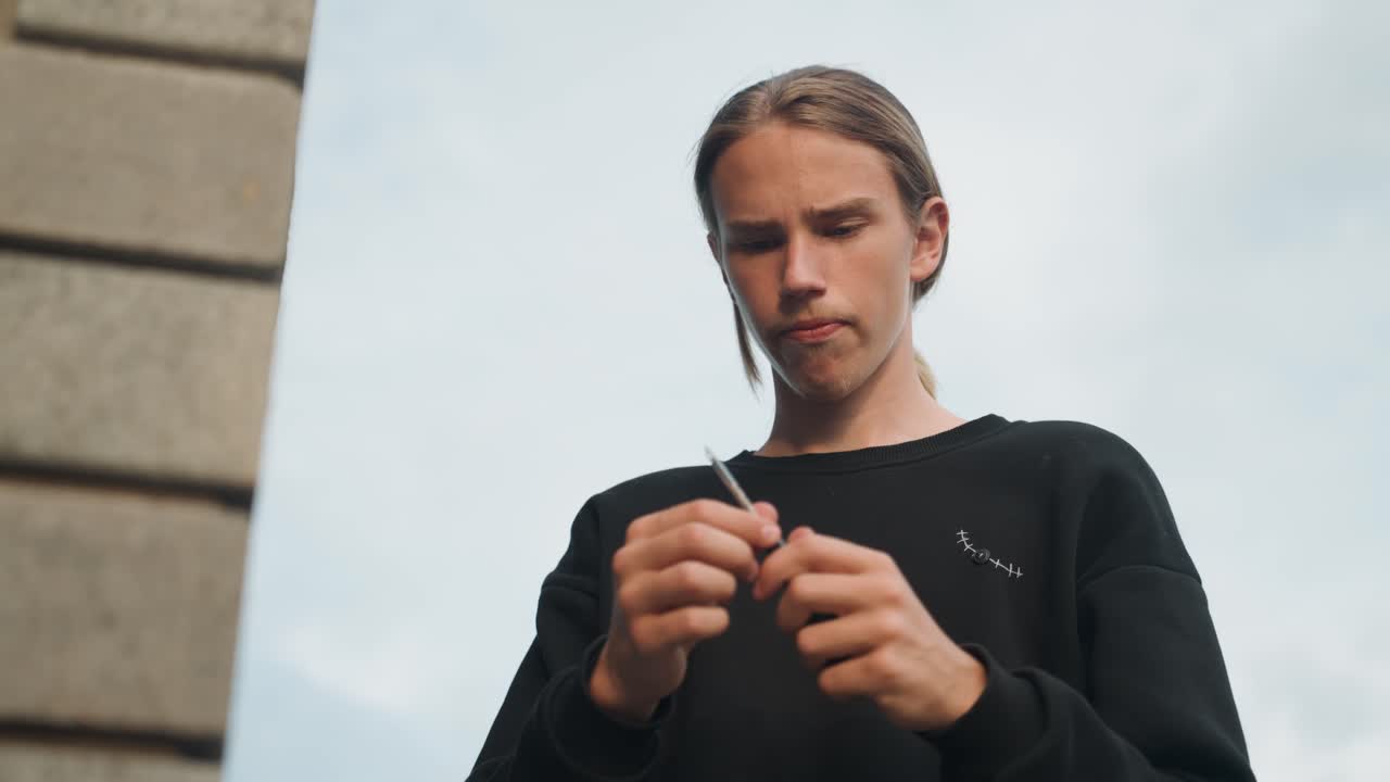 Ung mand forbereder en cigaret udendørs. En fokuseret person ruller en cigaret under en skyet himmel. En ung mand ruller og tænder forsigtigt en cigaret, mens et bymiljø omgiver ham.