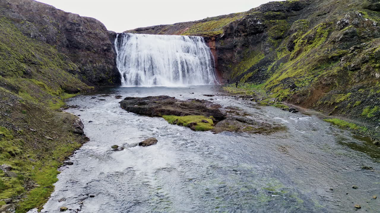 Aerial drone view of a large waterfall at the end of a river channel surrounded by moss covered cliffs and rocky slopes in Iceland's remote landscape
