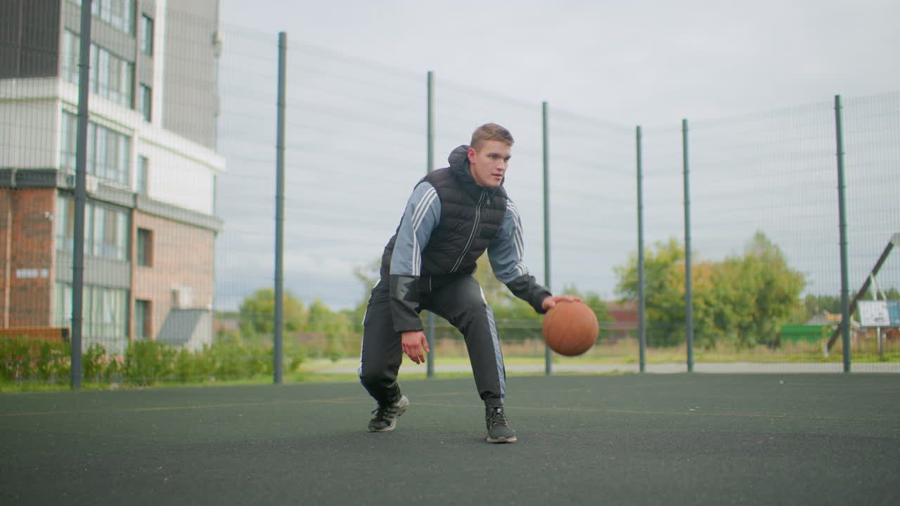 Atlet muda Kaukasia menggiring bola basket di luar ruangan di lapangan urban berpagar dekat gedung apartemen di bawah langit mendung, posisi bertahan rendah, ekspresi fokus, latihan solo dengan jaket
