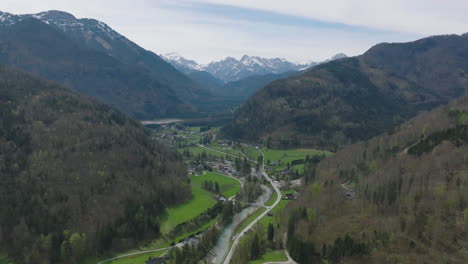 Vista-Aérea-Del-Pintoresco-Valle-Bajo-Las-Colinas-De-Los-Alpes-Austríacos,-Pueblo,-Río-Y-Carretera-En-Un-Paisaje-Verde