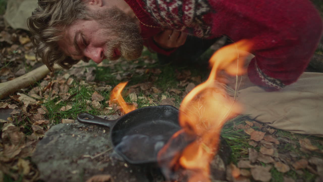 Man Bending to Forest Ground and Blowing into Campfire under Cooking Pan