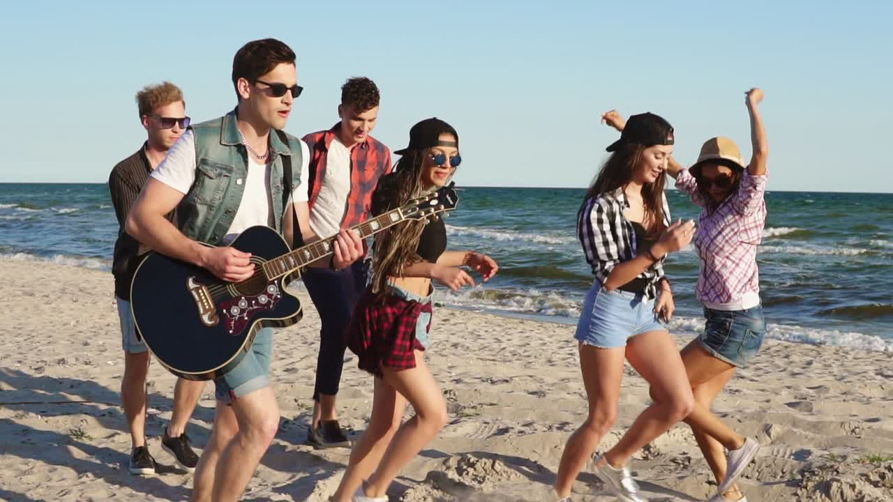 Hipster friends walking and dancing together playing guitar and singing songs on a beach at the water's edge. Slowmotion shot