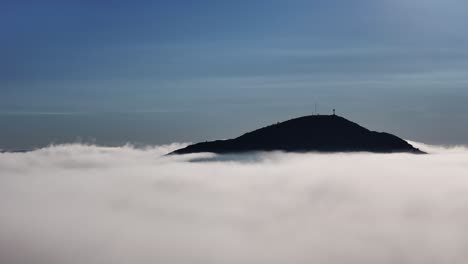 drone shot of cerro pan de azúcar in maldonado, uruguay. the mountain peak rises above low clouds as the intense sun breaks through, creating a dramatic and luminous scene
