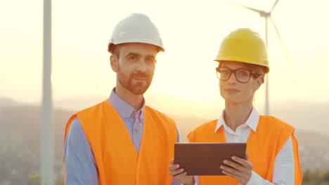 ingenieros caucásicos masculinos y femeninos con cascos y uniformes usando tabletas y hablando en la estación eólica de energía renovable, luego sonríen a la cámara