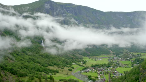 Majestuoso-Brote-Areal-Del-Paisaje-Del-Norte---Aurllandsfjord-flam-En-Noruega-Con-Un-Pequeño-Pueblo-En-La-Parte-Inferior-Derecha-Y-Una-Impresionante-Cascada-A-La-Izquierda-Y-Un-Dron-Que-Se-Acerca-Lentamente-A-La-Cascada